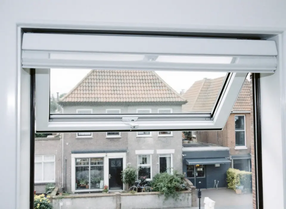 Een open klepraam boven een woonkamerraam, met zicht op bakstenen huizen aan de overkant van de straat